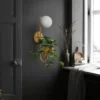 Minimalist Warm Glass Plant Sconce with a frosted globe light and hanging greenery, mounted on a dark wall beside a bright window.