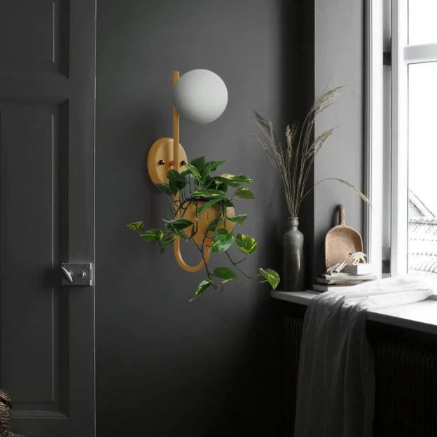 Minimalist Warm Glass Plant Sconce with a frosted globe light and hanging greenery, mounted on a dark wall beside a bright window.
