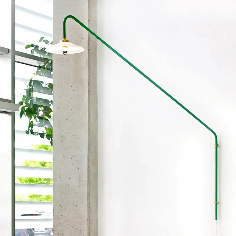N3 Hanging Wall Lamp with a sleek green arm and floating shade, casting soft light against a clean wall near a window with greenery.