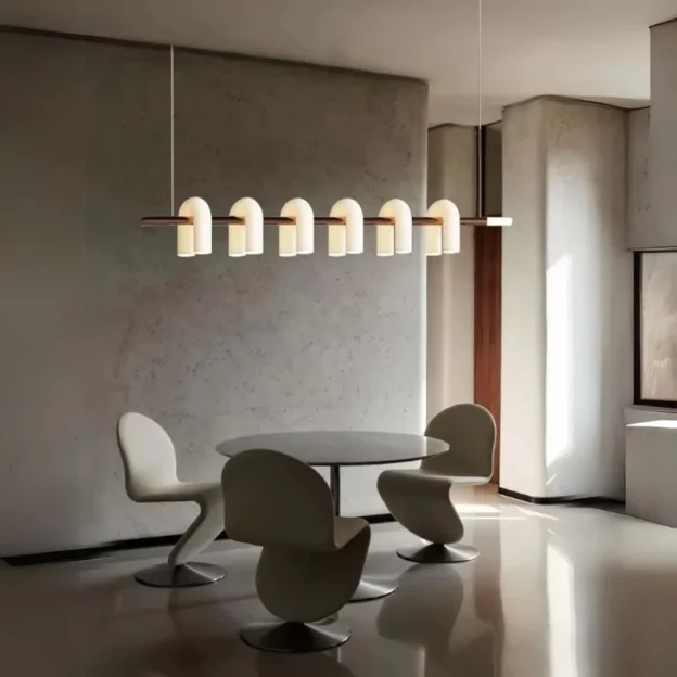 Minimalist dining room with round table and sculptural chairs lit by the Odyssey Linear Chandelier featuring curved warm-glow diffusers.