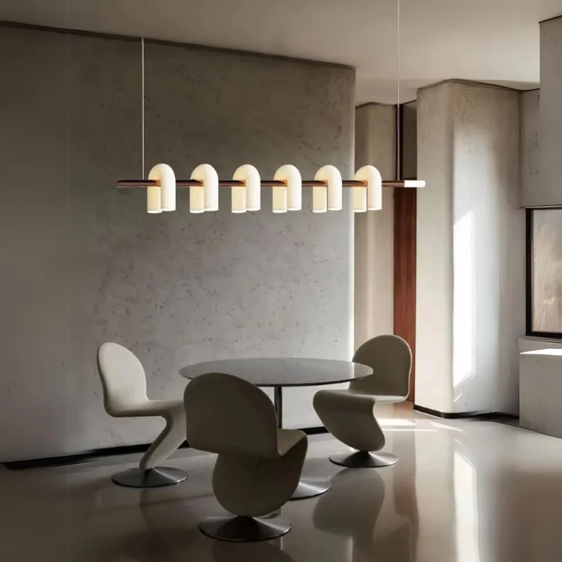 Minimalist dining room with round table and sculptural chairs lit by the Odyssey Linear Chandelier featuring curved warm-glow diffusers.