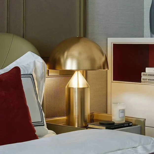 The Metal Atollo Table Lamp with a sleek golden dome design, placed on a bedside table with books and a candle in a cozy bedroom.