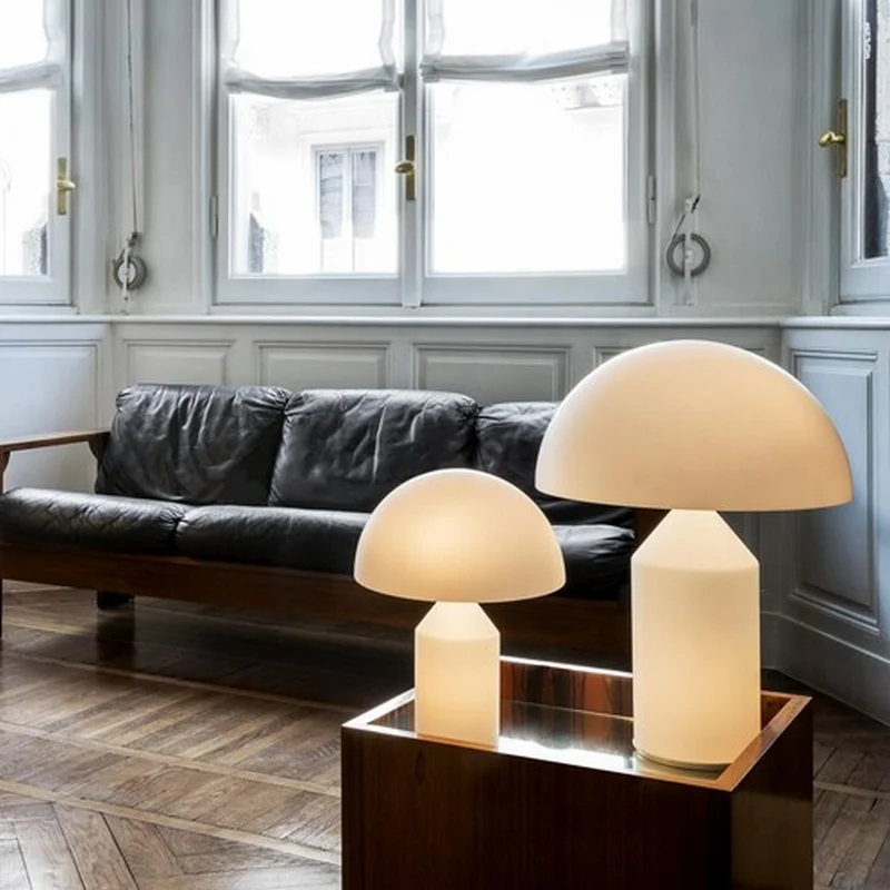 The Glass Atollo Table Lamp features a sleek mushroom-shaped design with a soft, glowing light, placed beside a leather sofa.